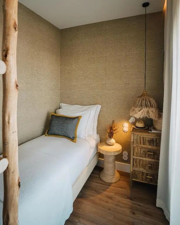 Schmaler Schlafbereich mit Einzelbett, beigefarbener Tapete und Nachttisch aus Holz.