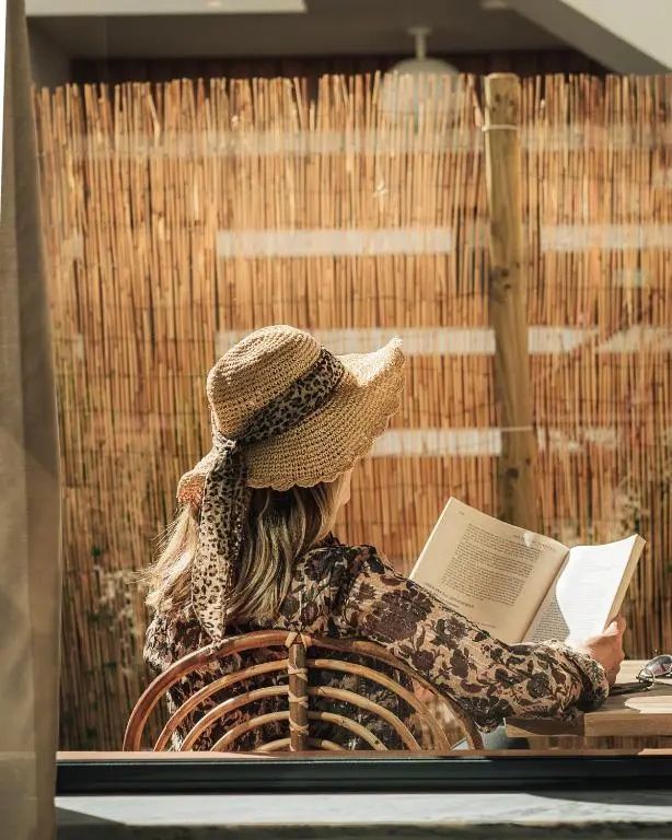 Frau mit Sonnenhut liest entspannt ein Buch auf einem Flechtstuhl im Außenbereich.