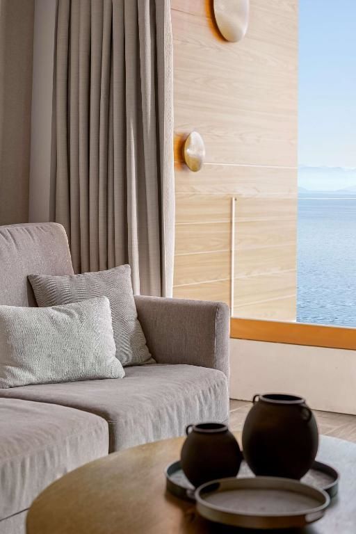 Sitzbereich mit beigem Sofa und Kissen vor einer Holzwand mit Blick auf das blaue Meer.