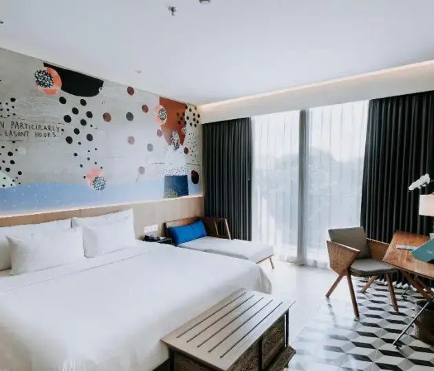 Modernes Hotelzimmer mit Doppelbett, Wandgemälde, Tagesbett und Sitzbereich am Fenster.