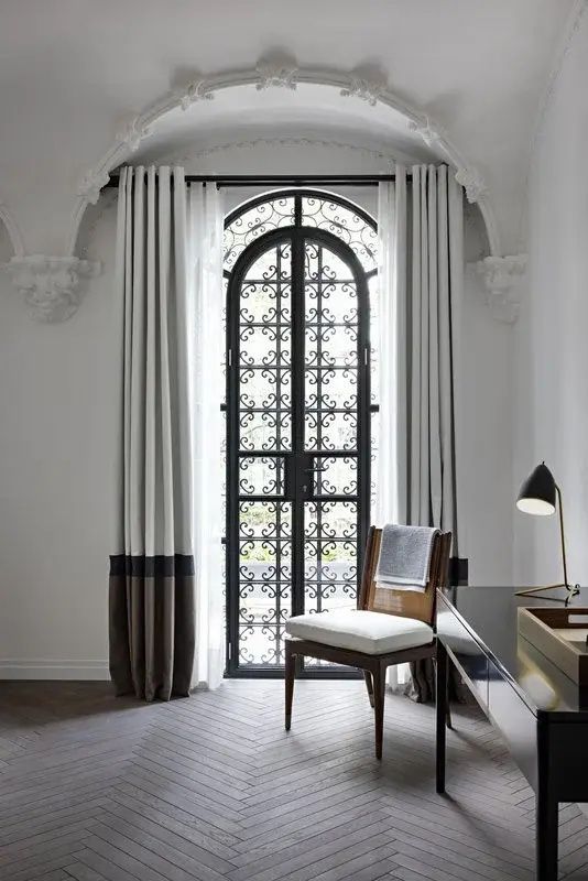 Schreibtisch mit Lampe und Stuhl vor einem hohen Bogenfenster mit schwarzem Gitter.