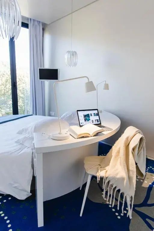 Helles Hotelzimmer mit weiß bezogenem Bett, integriertem Schreibtisch, Stuhl und Blick ins Grüne.