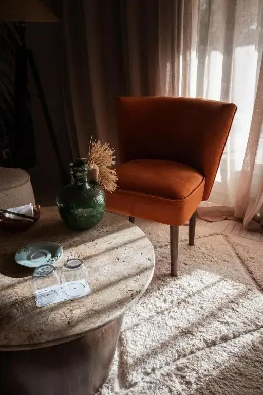 Oranger Sessel neben einem runden Steintisch mit grüner Vase und Gläsern im Sonnenlicht.