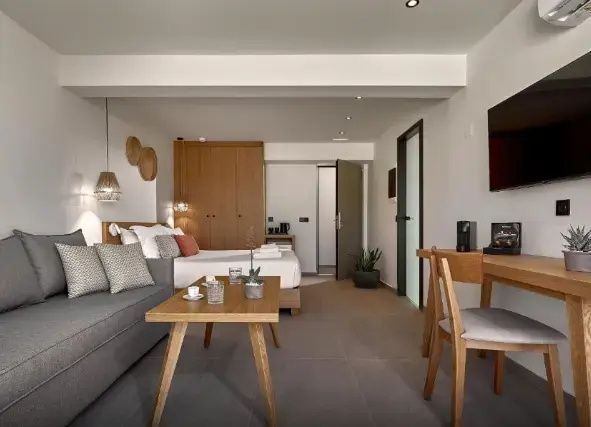 Helles Hotelzimmer mit Holztisch im Vordergrund, Sofa und Doppelbett im Hintergrund.