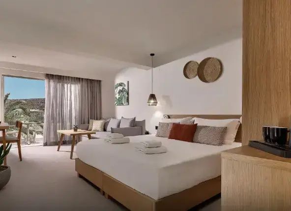 Doppelbett mit braunen Kissen neben einem Sofa mit Holztisch und Vorhängen am Fenster.