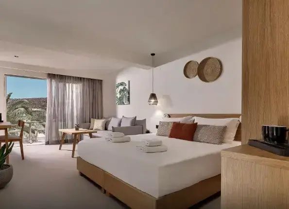Helles Hotelzimmer mit Doppelbett, Sitzsofa, Holztisch und Zugang zum Balkon.
