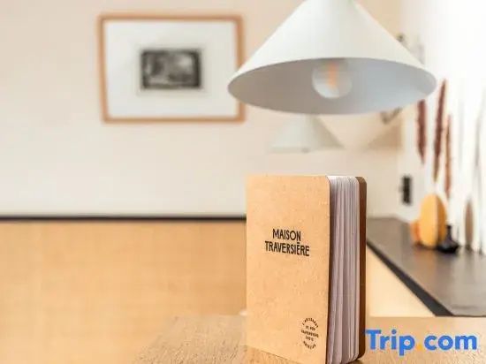 Kleine Broschüre mit Aufschrift Maison Traversière auf einem Holzmöbel unter einer Lampe.