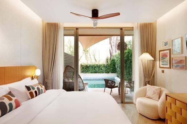 Helles Hotelzimmer mit Doppelbett, Sessel und Blick durch die Glasfront auf eine Terrasse mit Pool.