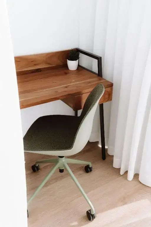 Schreibtisch aus Holz mit einem grünen Bürostuhl in einer Nische neben weißen Vorhängen.
