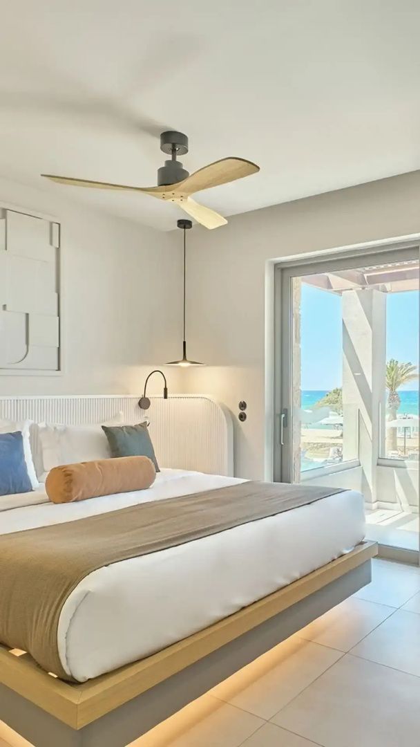 Hell eingerichtetes Doppelzimmer mit Ventilator und Blick durch die Balkontür auf das Meer.