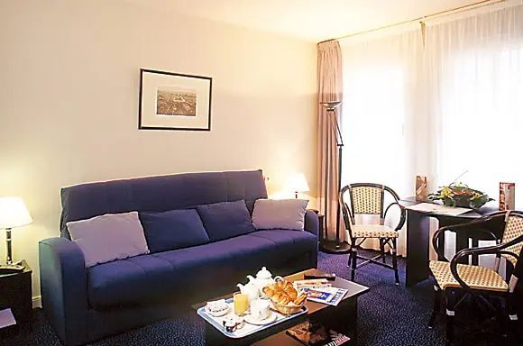 Wohnzimmer mit blauem Sofa, Beistelltisch, Sessel und hellem Vorhang.