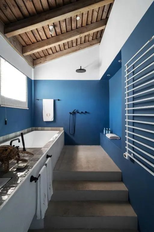 Badezimmer mit blauer Wandgestaltung, eingelassener Badewanne und offener Dusche unter Holzgebälk.