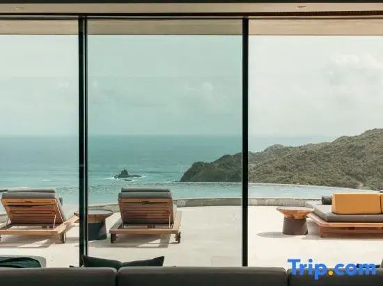 Blick durch ein Fenster auf einen Infinity-Pool, das Meer und grüne Hügel.