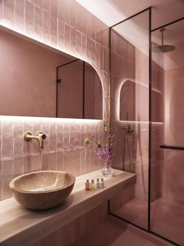 Badezimmer mit rosa gefliesten Wänden, einem Waschtisch aus Stein, beleuchtetem Spiegel und Dusche.
