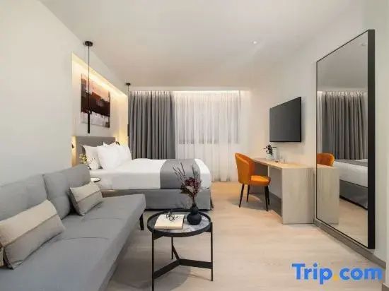 Doppelbett mit grauer Bettwäsche, Sofa und Schreibtisch in einem hellen Hotelzimmer.