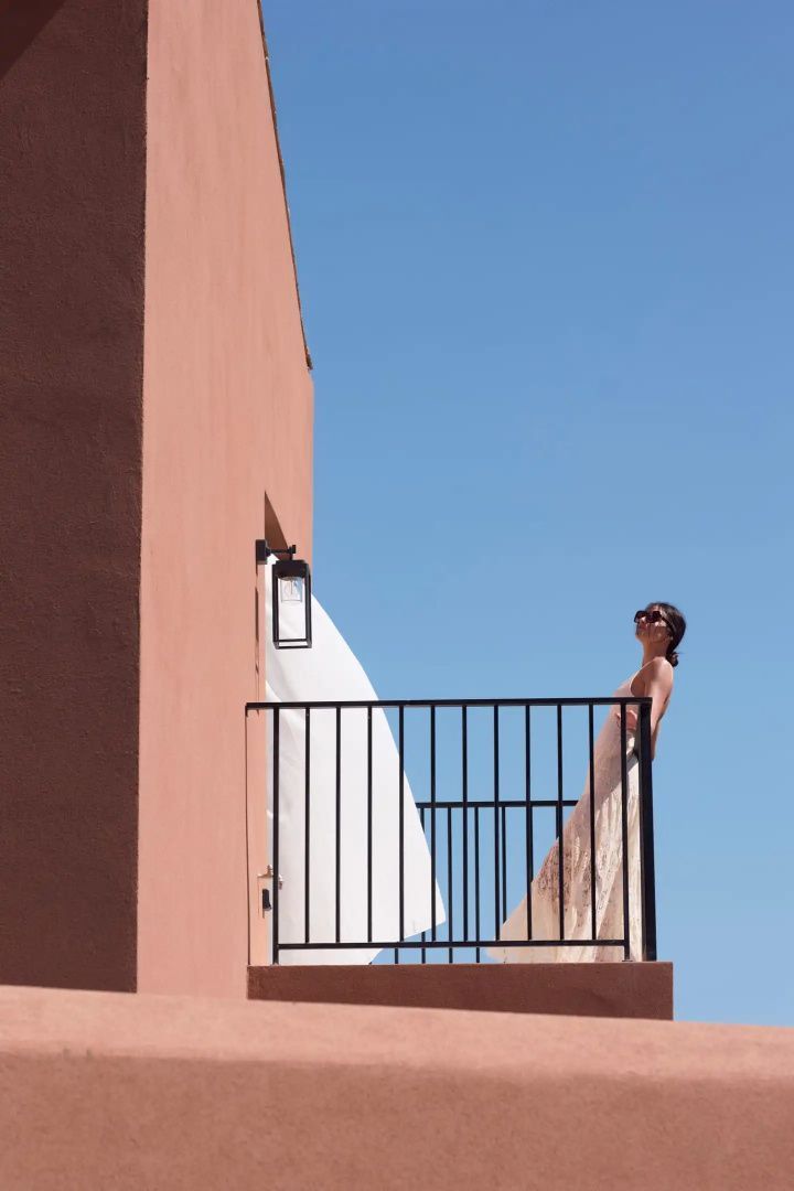 Eine Frau steht auf dem Balkon eines terracottafarbenen Gebäudes unter blauem Himmel.