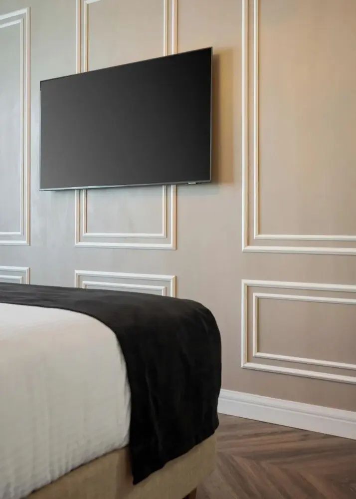An einer hellen Wand mit Zierleisten hängt ein Flachbildfernseher über einem Bett.