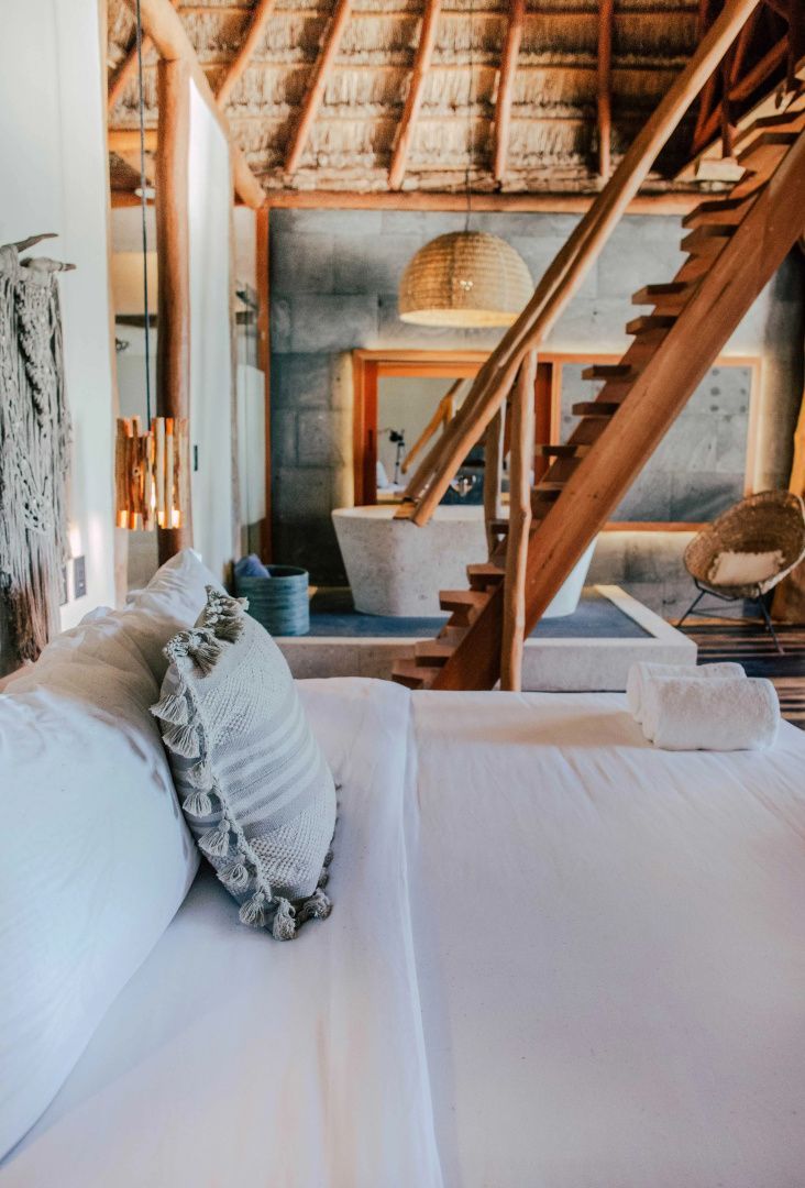 Schlafbereich mit weiß bezogenem Bett, Holztreppe und Badewanne im Hintergrund.