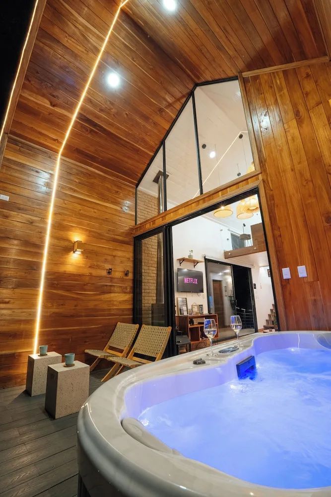 Holzterrasse mit eingelassenem Whirlpool, zwei Liegestühlen und Blick in das beleuchtete Innere der Hütte.