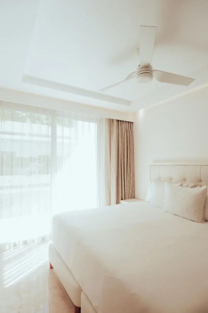 Helles Schlafzimmer mit weißem Doppelbett, Deckenventilator und großem Fenster zum Balkon.