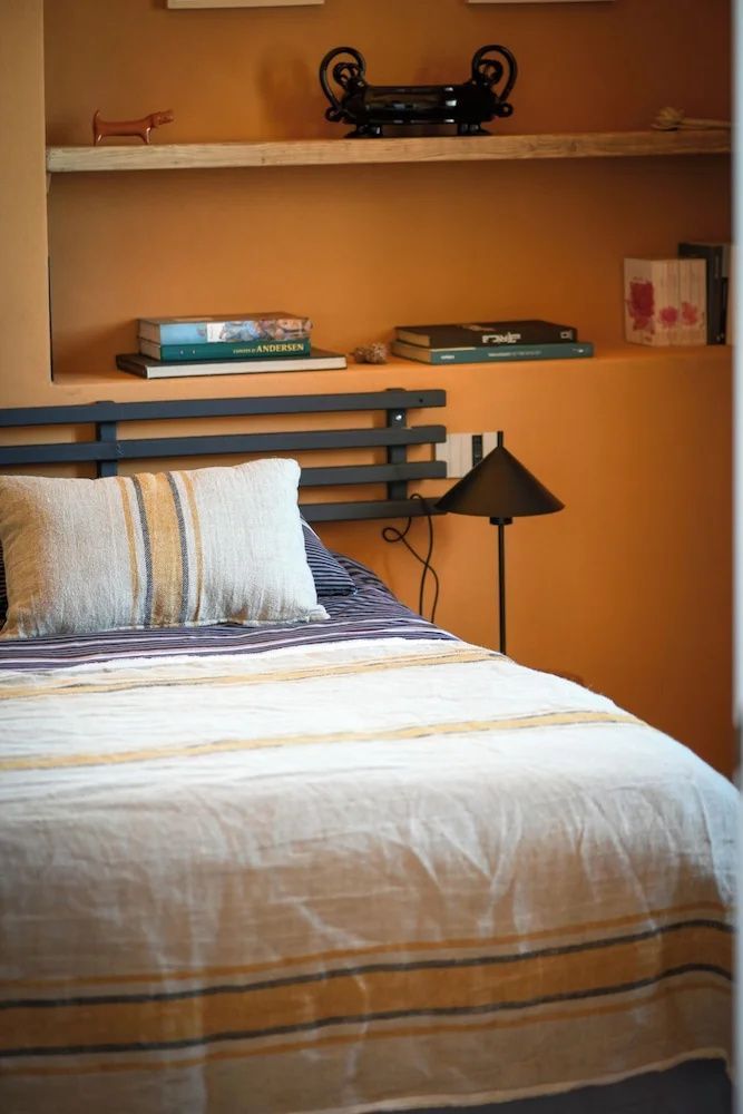 Bett mit gestreiftem Kissen, dunklem Kopfteil und Leselampe neben einer Nische mit Büchern.