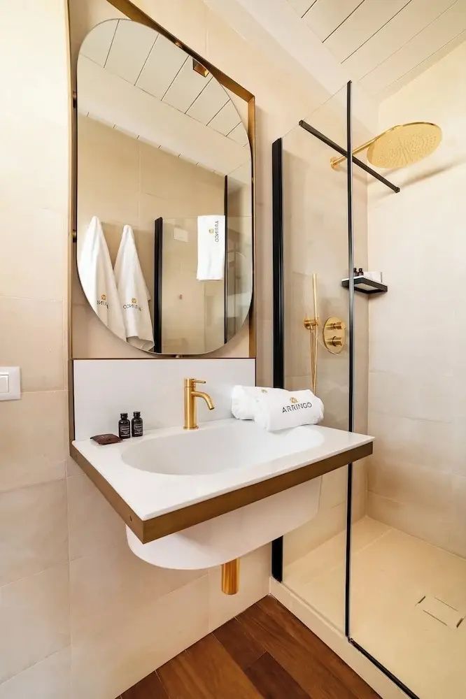 Waschtisch mit goldener Armatur, ovalem Spiegel und angrenzender Glasdusche.