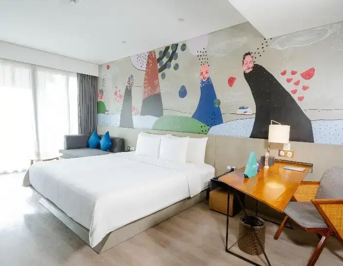 Hell eingerichtetes Hotelzimmer mit einem Doppelbett, bunten Kunstwerken an der Wand und einem Schreibtisch.