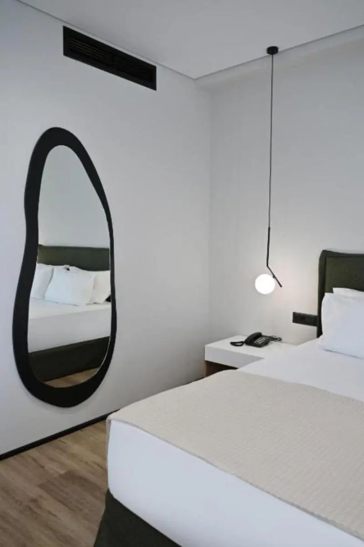 Doppelbett mit weißer Bettwäsche, Nachttisch und organischer Wandspiegel im Hotelzimmer.