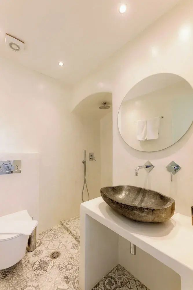 Modernes Badezimmer mit Steinwaschbecken, rundem Spiegel, Toilette und einer begehbaren Dusche.