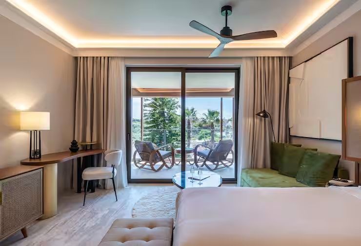 Hotelzimmer mit Schreibtisch, Deckenventilator und Glasbalkontür mit Blick ins Grüne.