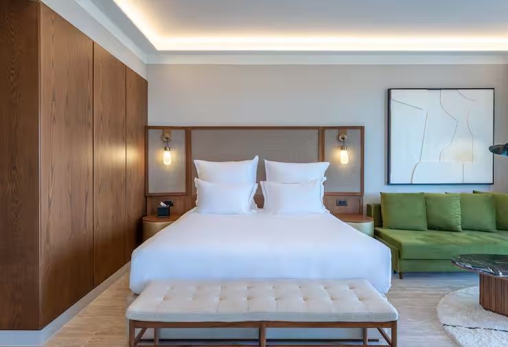 Helles Doppelbett mit weißer Bettwäsche, Kopfteil aus Holz, Sitzbank und grünem Sofa.