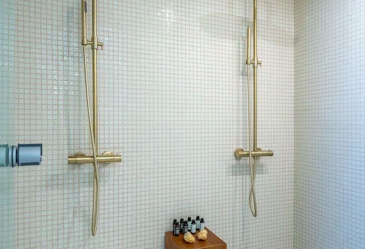 Zwei goldene Duscharmaturen an einer hell gefliesten Wand mit kleinen Mosaikfliesen.