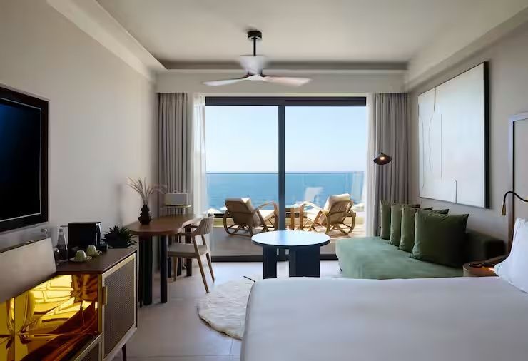Helles Hotelzimmer mit grünem Sofa, Holztisch und Blick auf die Terrasse mit Meerblick.