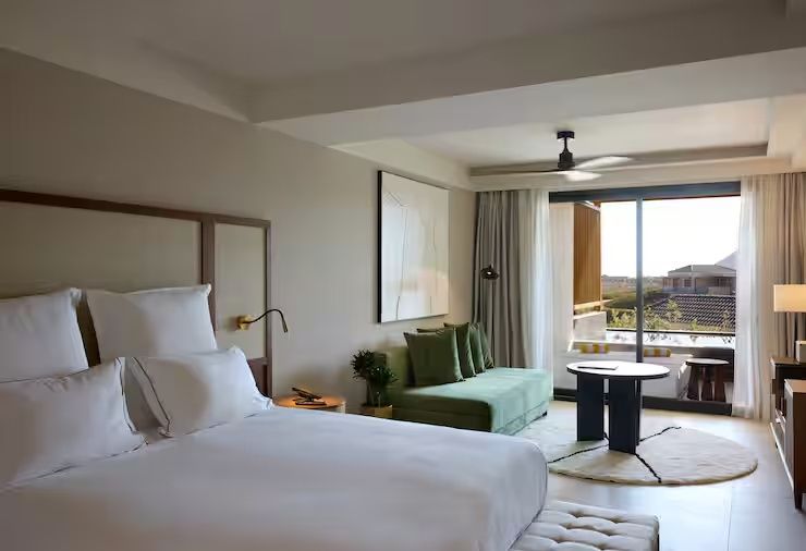Moderne Suite mit weißem Bett, grünem Sofa und großen Fenstern zum sonnigen Balkon.