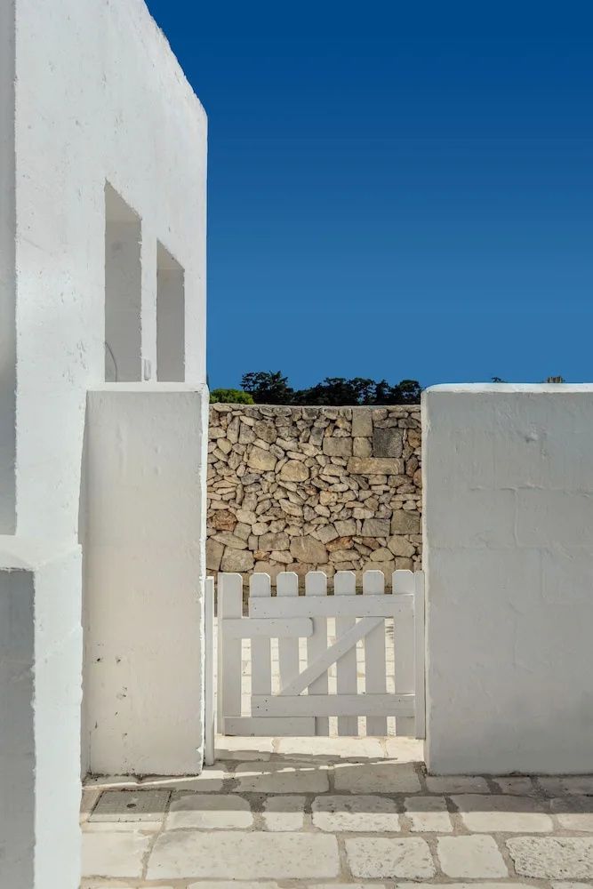 Weiße Gebäudeaußenwand mit kleinem Tor vor einer traditionellen Steinmauer.