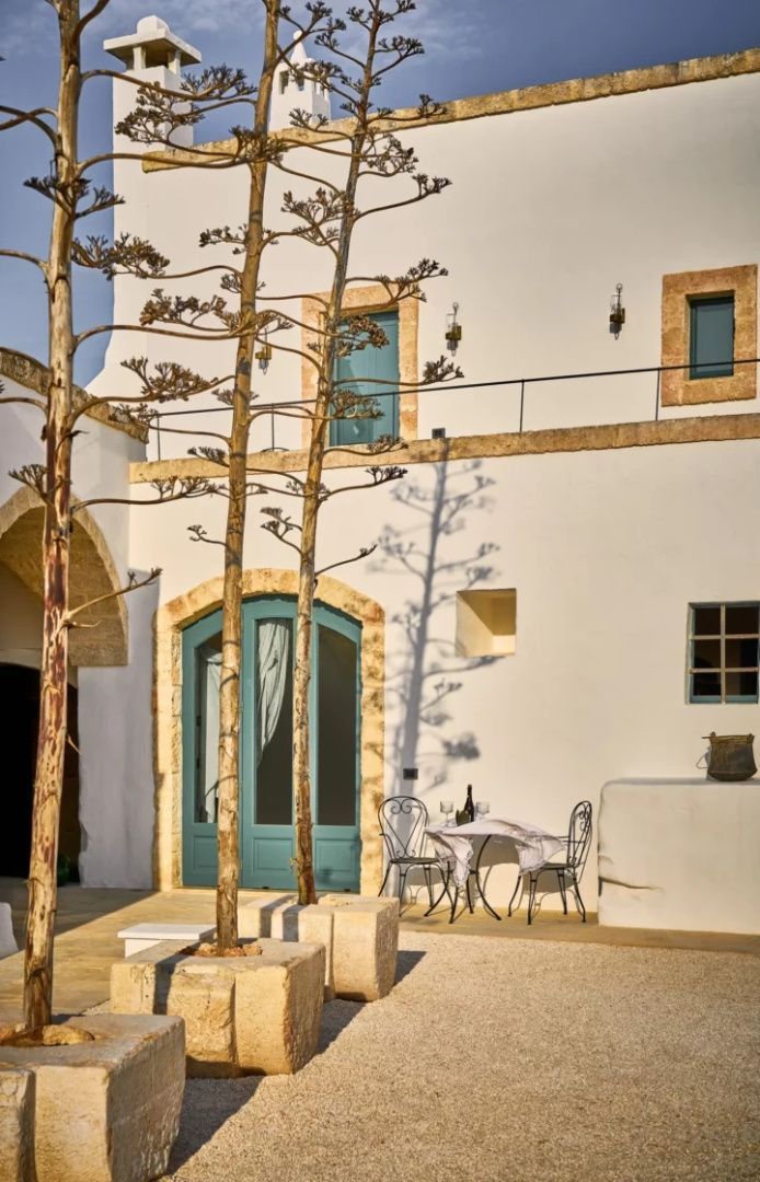 Weiße Fassade der Masseria mit türkisfarbenen Türen, hohen Agaven und einer kleinen Sitzgruppe im Außenbereich.