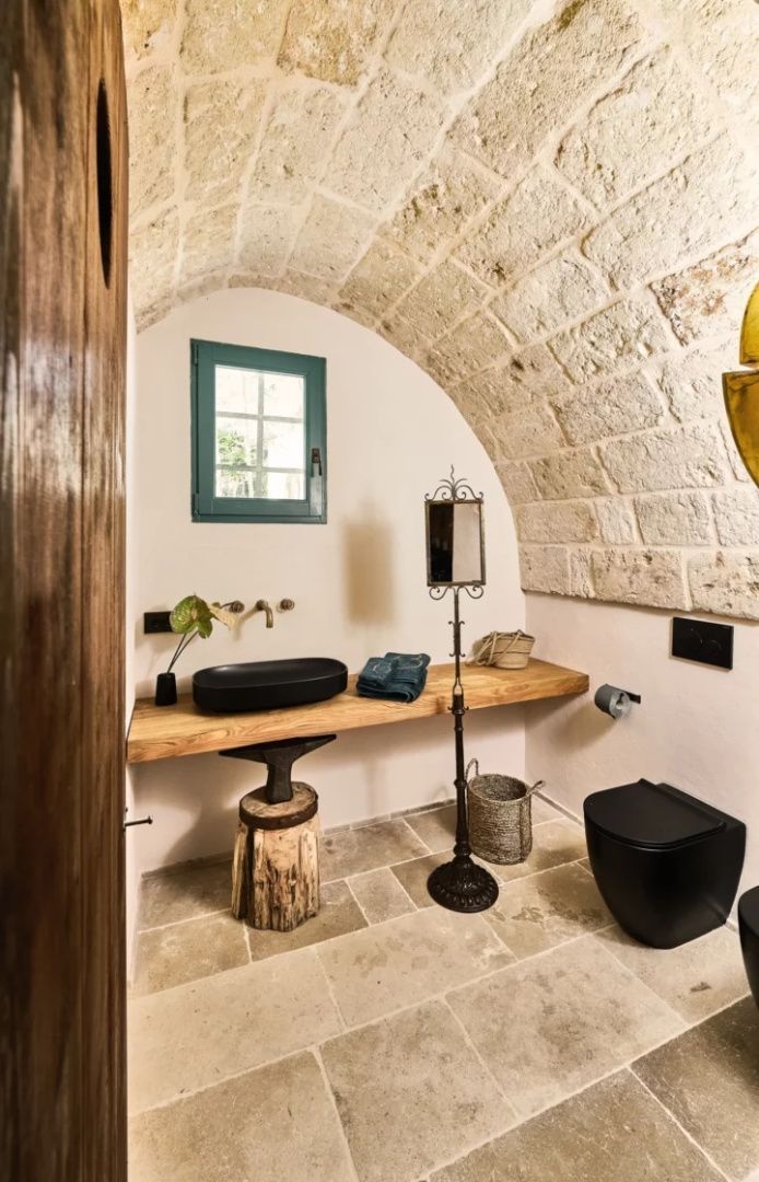 Badezimmer mit gewölbter Steinwand, Holzwaschtisch, schwarzem Waschbecken und WC.