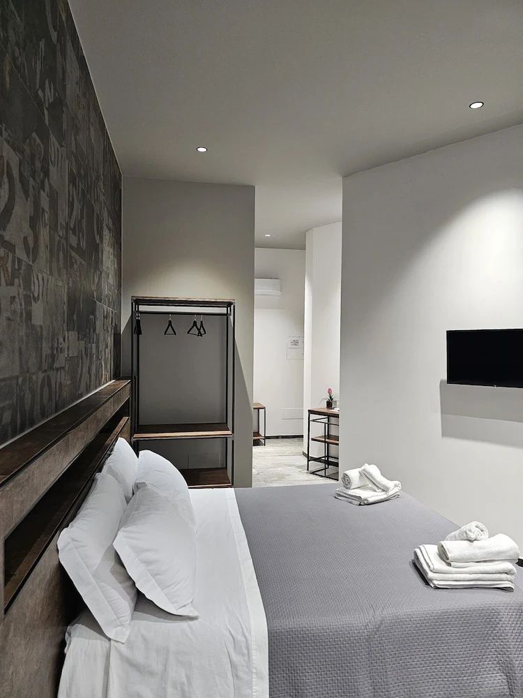 Doppelbett mit grauer Decke, weiße Handtücher, Wandfernseher und offener Kleiderschrank.