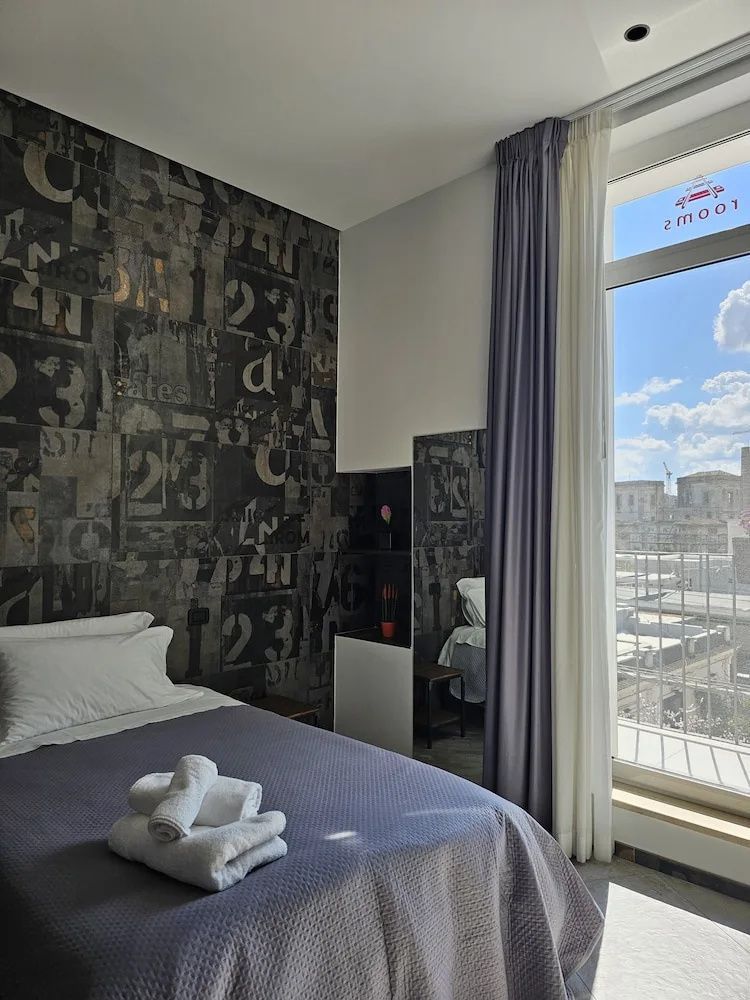 Doppelbett mit Handtüchern im Hotelzimmer mit gemusterter Tapete und hohem Fenster.