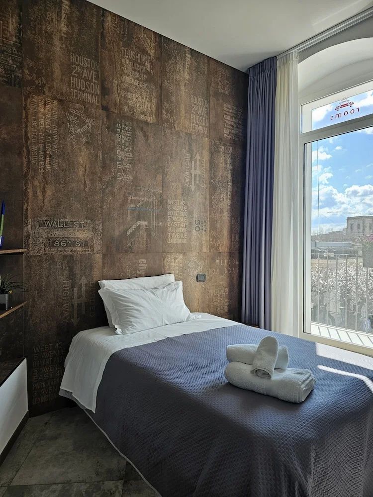 Bett mit weißem Kissen vor braun gemusterter Wand neben einem hellen Fenster.
