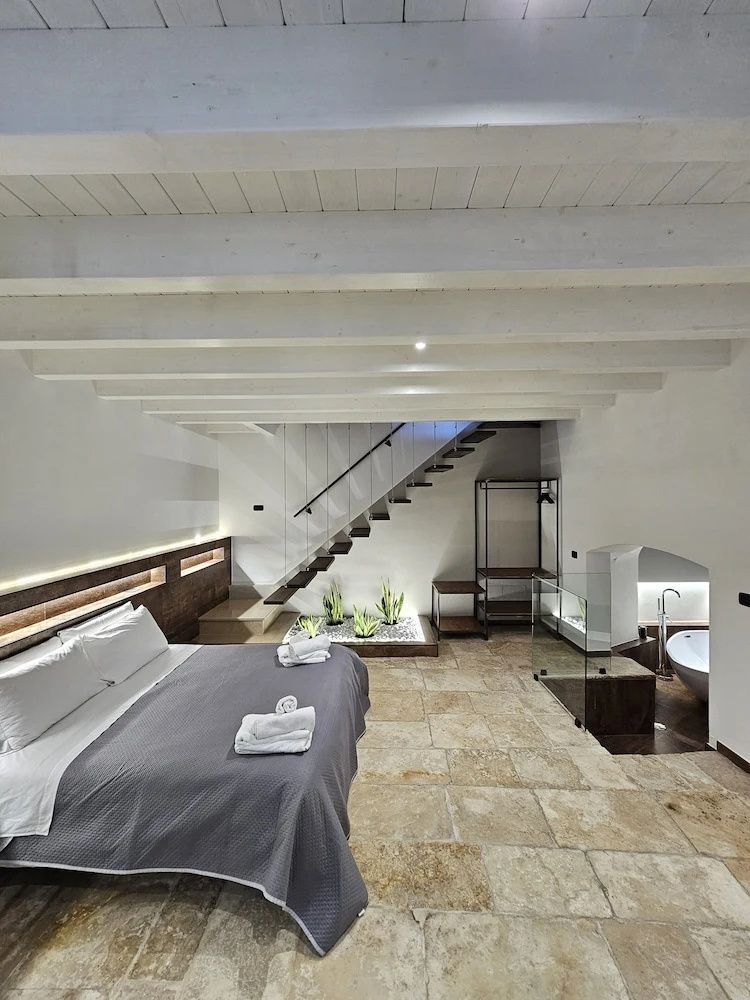 Doppelbett mit grauer Tagesdecke, Treppe ins Obergeschoss und sichtbare Holzbalken an der Decke.