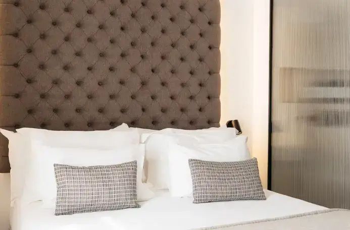 Braunes gepolstertes Kopfteil mit Knöpfen hinter einem weiß bezogenen Doppelbett mit Kissen.