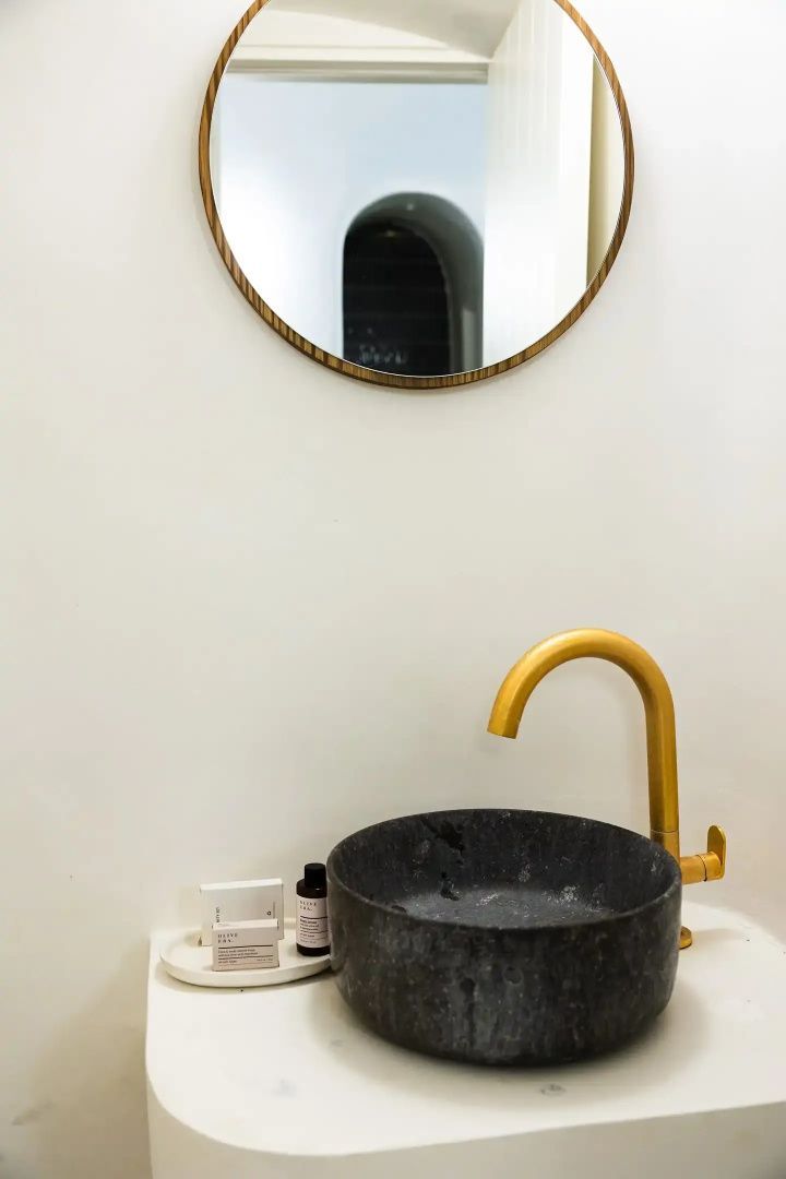 Dunkles Steinwaschbecken mit goldener Armatur und rundem Spiegel an weißer Wand