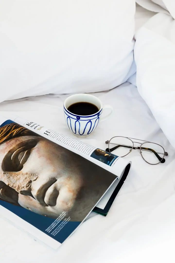 Eine Kaffeetasse, eine Brille und ein Magazin liegen auf weißem Bettzeug