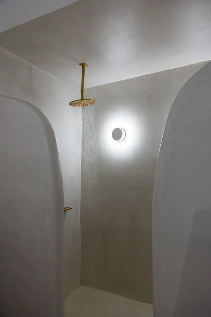 Regendusche mit goldener Armatur an einer hellen Wand mit runder Wandleuchte.