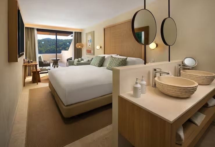 Hell eingerichtetes Hotelzimmer mit Doppelbett, Waschtisch im Vordergrund und Zugang zum Balkon.