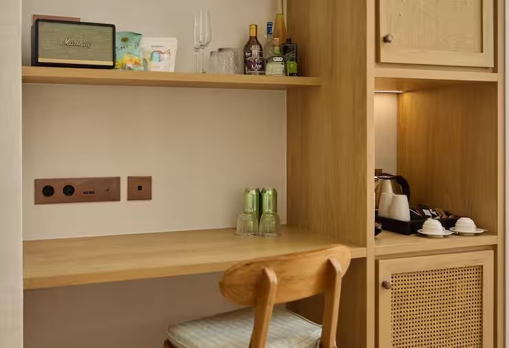 Holzschreibtisch und Regalelement mit Minibar, Gläsern, Wasserkocher und Stuhl.