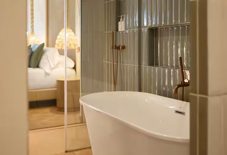 Freistehende weiße Badewanne vor grauen Fliesen mit Blick ins Schlafzimmer.