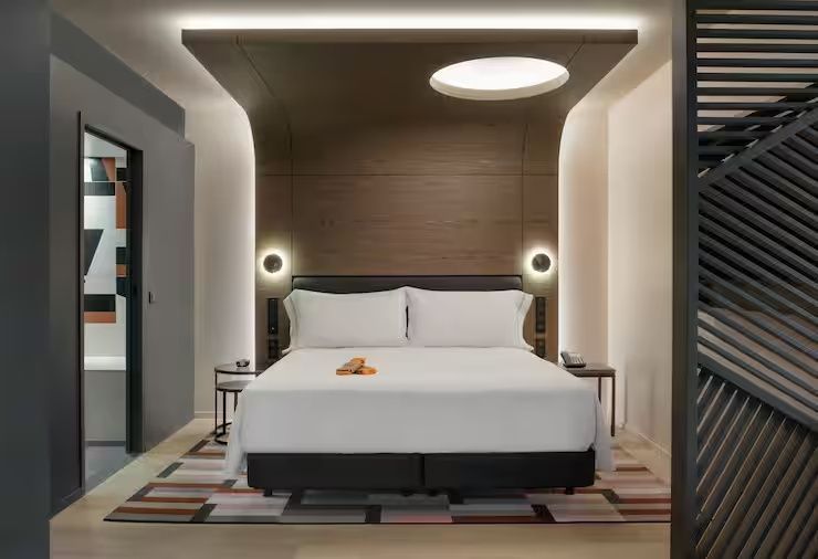 Doppelbett im Hotelzimmer mit Holz-Baldachin, indirekter Beleuchtung und buntem Teppich.