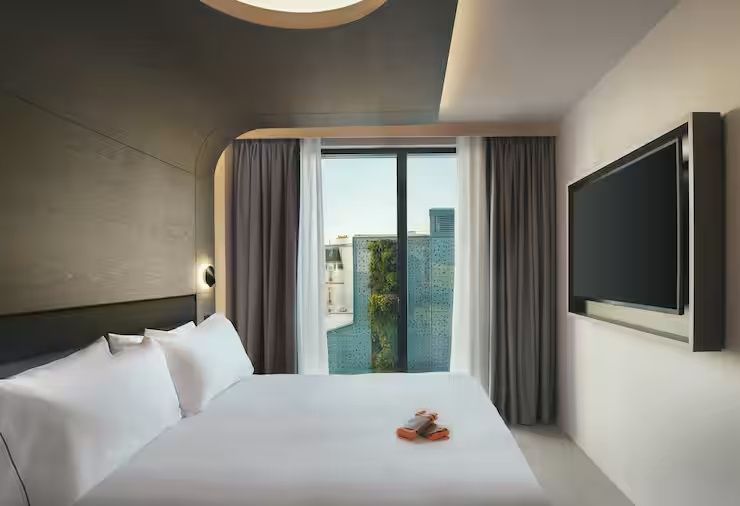 Modernes Hotelzimmer mit weiß bezogenem Doppelbett, Wandfernseher und Blick ins Grüne durch das Fenster.